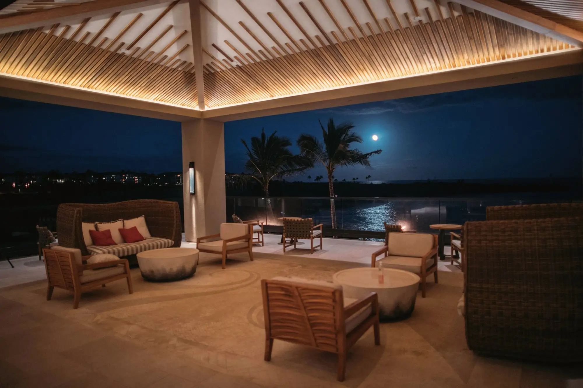 An open-air resort lounge at night, warm light under a timber ceiling and a full moon over the water