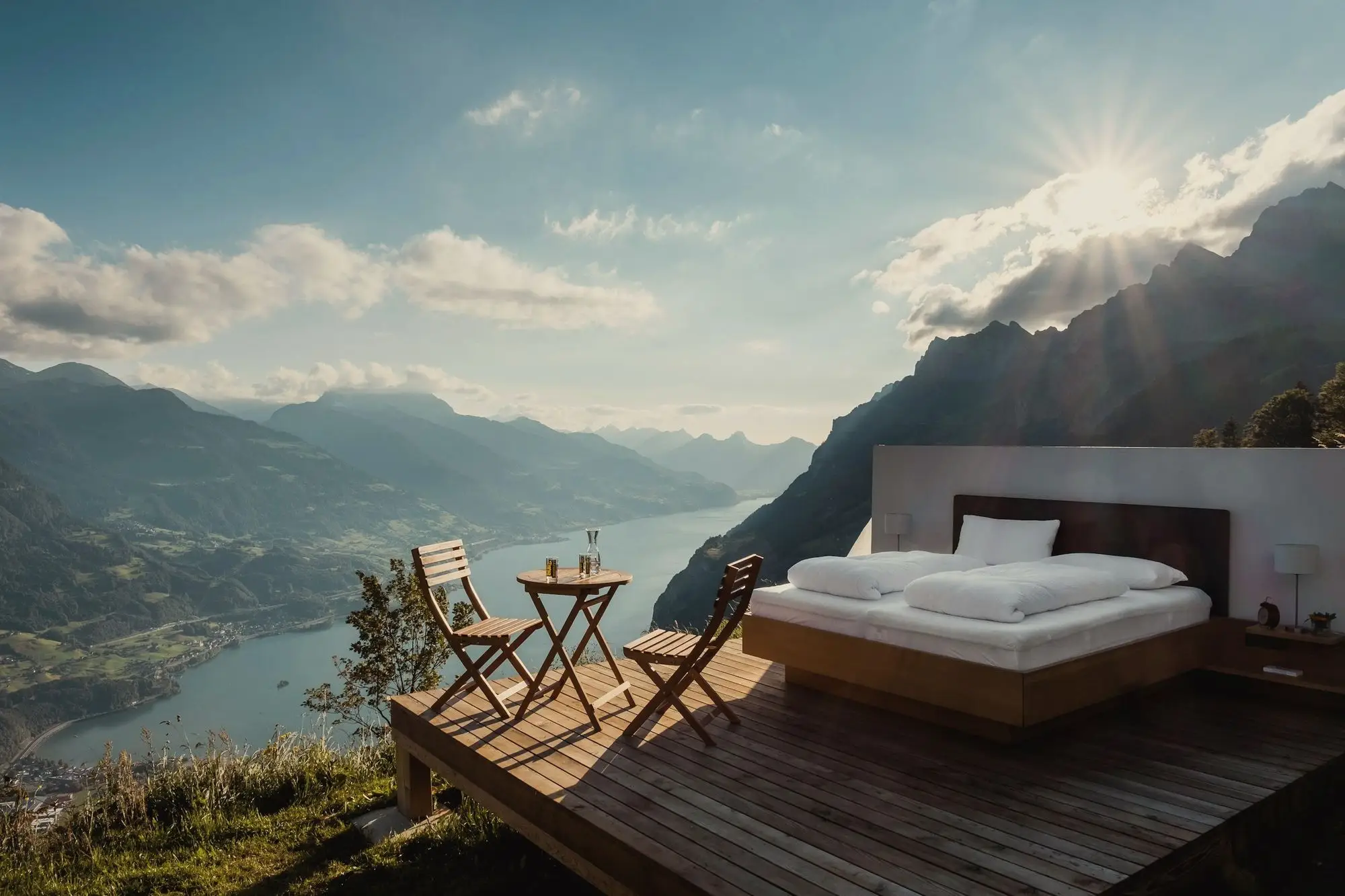 A bed on an open-air deck high above a mountain lake, lit by the morning sun